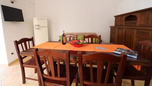 Uma área de refeições com uma mesa, cadeiras, uma tigela de frutas, uma garrafa de vinho e uma televisão na parede.