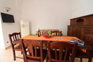 Ein Essbereich mit Tisch, Stühlen, einer Obstschale, einer Weinflasche und einem Fernseher an der Wand.