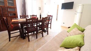 Uma sala de jantar com uma mesa e cadeiras de madeira, uma televisão fixada na parede e um sofá com almofadas verdes.