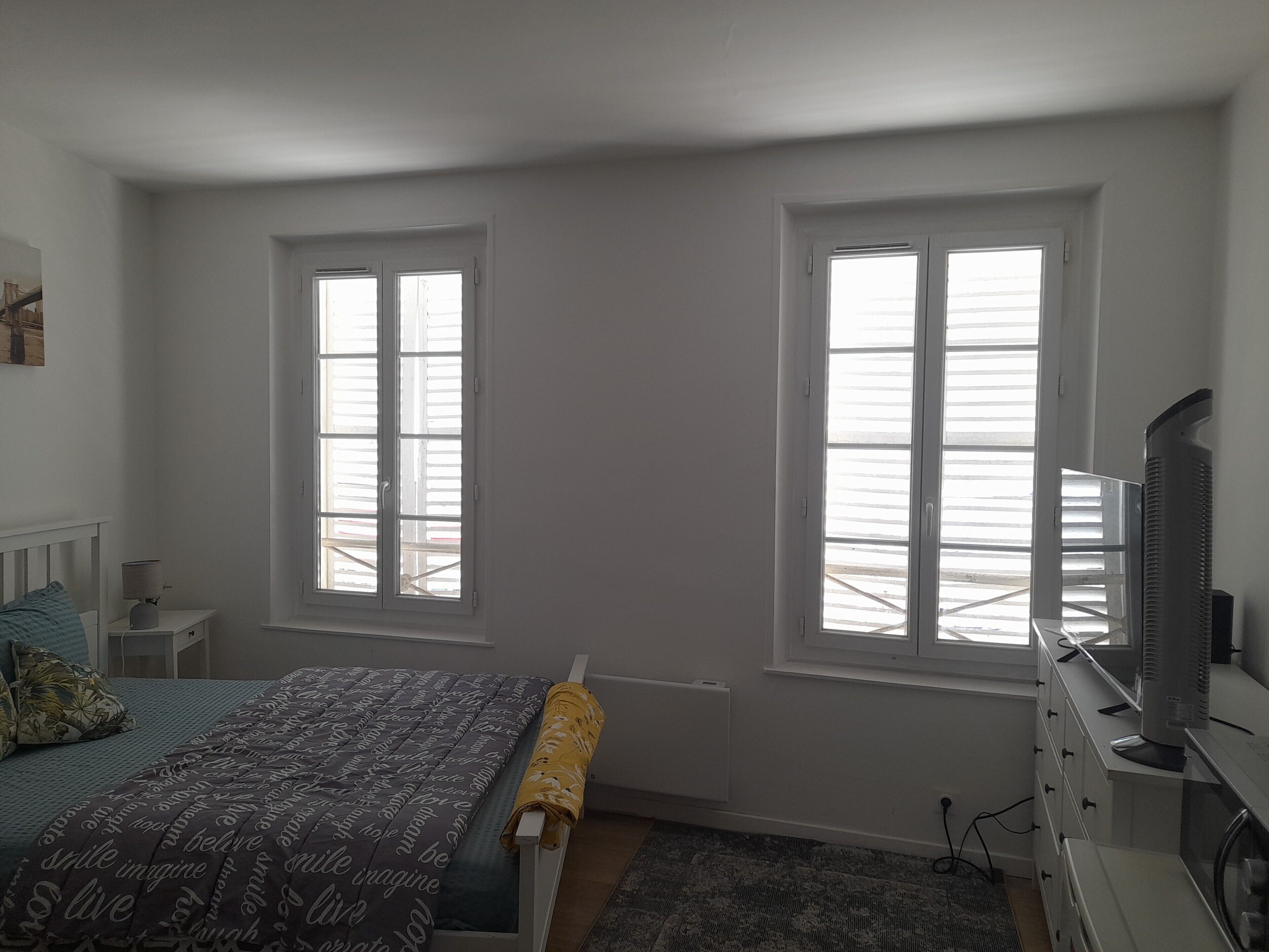 Un dormitorio con una cama, dos ventanas, un aparador y un televisor.
