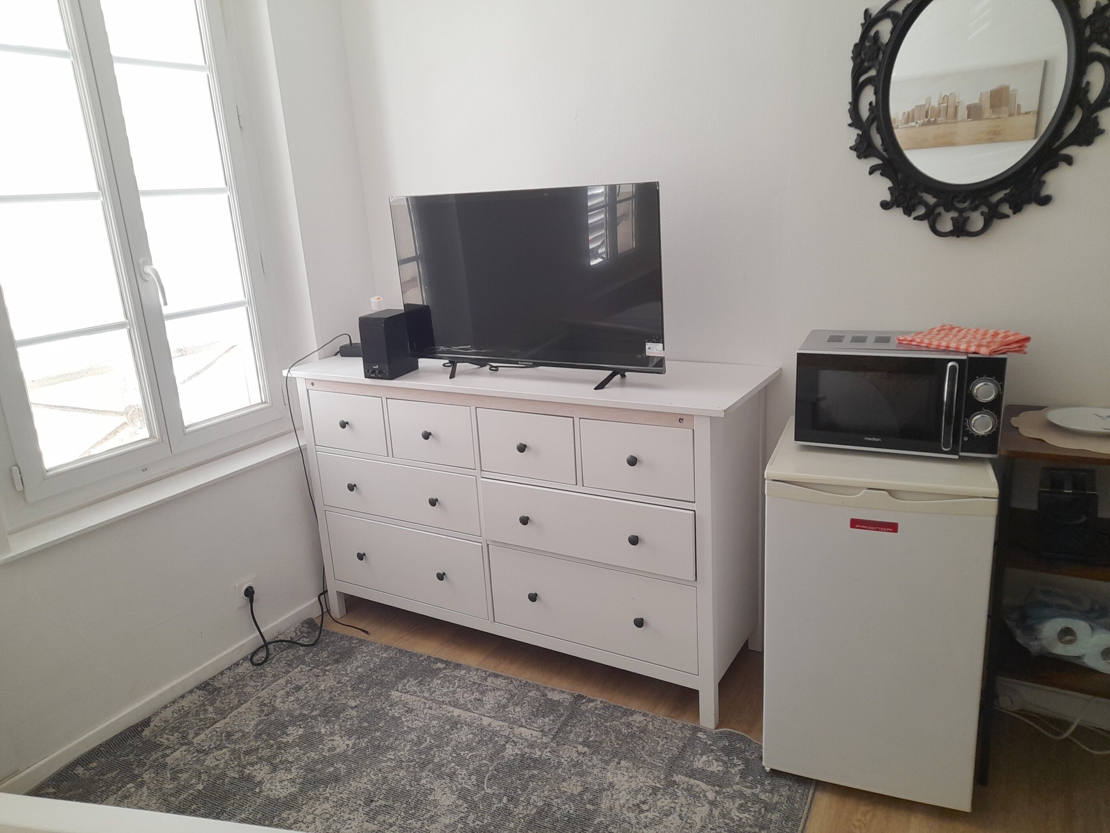 Een kamer met een witte ladekast, een flatscreen televisie, een magnetron en een kleine koelkast.