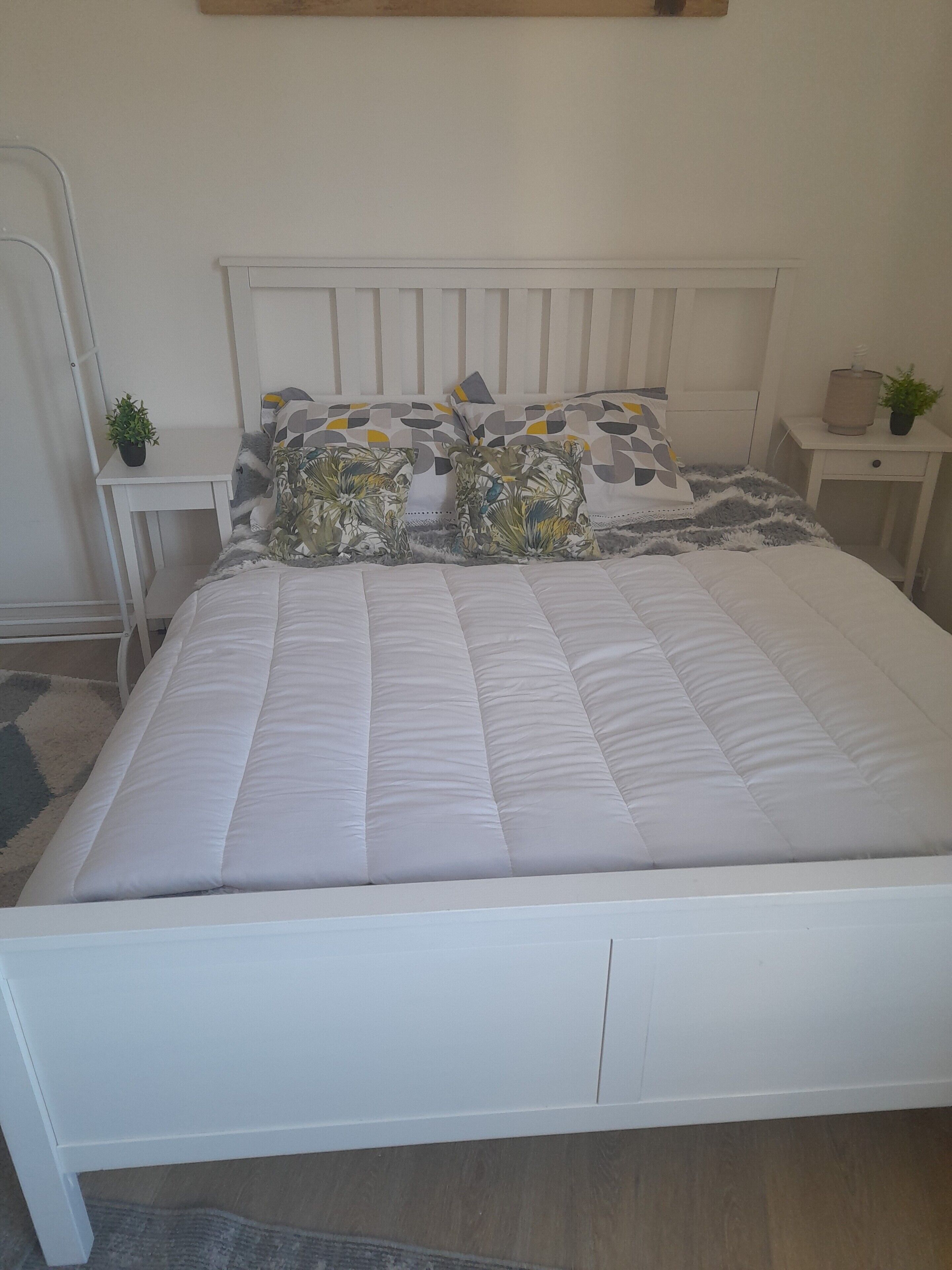 Een wit bed met een gewatteerde matras, een houten hoofdbord en een nachtkastje met een plant.
