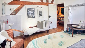 Habitació amb sostre de fusta, un llit amb colcha floral, una cadira amb coixí quadriculat, taula petita amb un plat de llaminatures i un bany amb rentapèus i mirall.