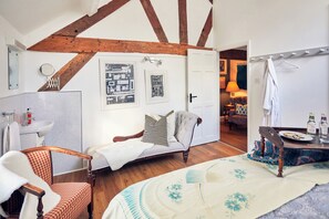 A room with a wooden ceiling, a bed with a floral bedspread, a chair with a checkered cushion, a small table with a plate of snacks, and a bathroom with a sink and mirror. - Apple Bough Cottage (Hereford)