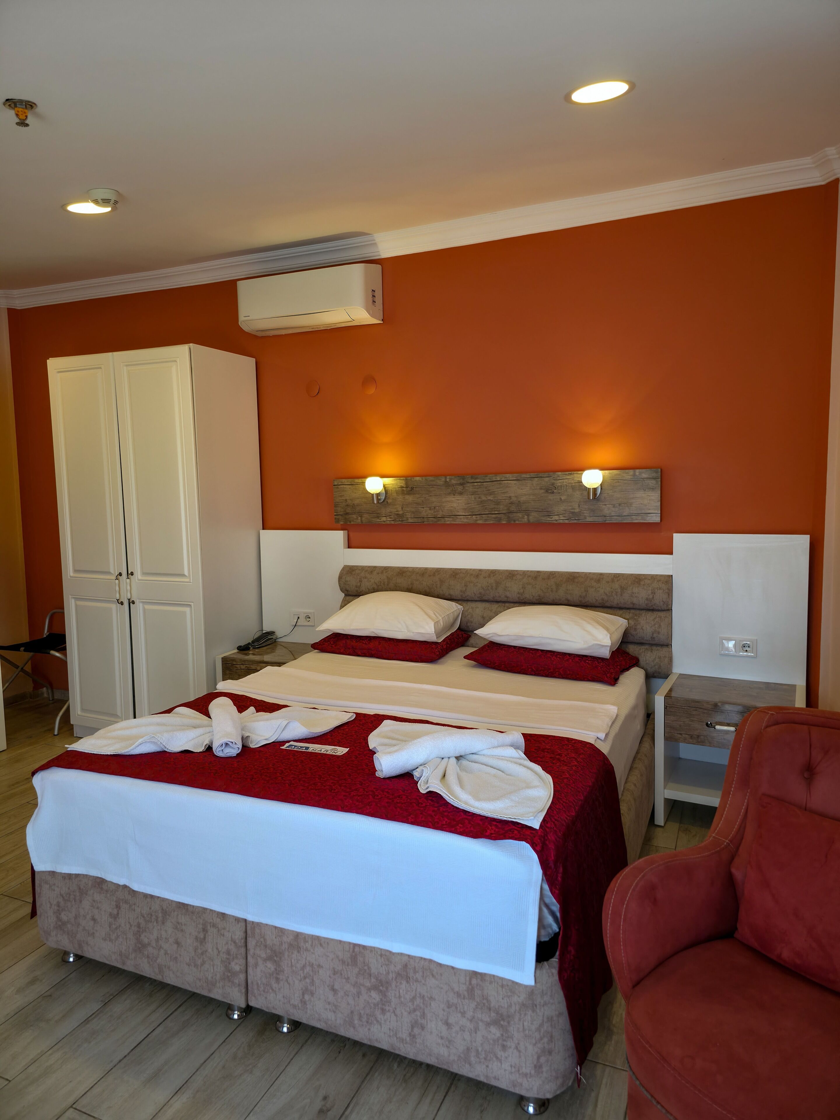 Una cama doble con colcha roja y blanca, dos mesitas de noche, un armario y un sillón rojo.