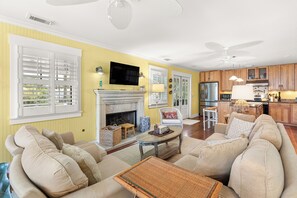 A living room with a fireplace, a television, and a kitchen area with wooden cabinets and a refrigerator. - Private Heated Pool, Rooftop Views & Steps to Best Beaches by Tybee Cottages (Tybee Island)