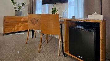 A room with a television mounted on the wall, a wooden desk with a computer, a chair, and a small refrigerator.