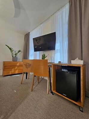 Una habitación con televisión instalada en la pared, un escritorio de madera con computadora, una silla y un refrigerador pequeño.