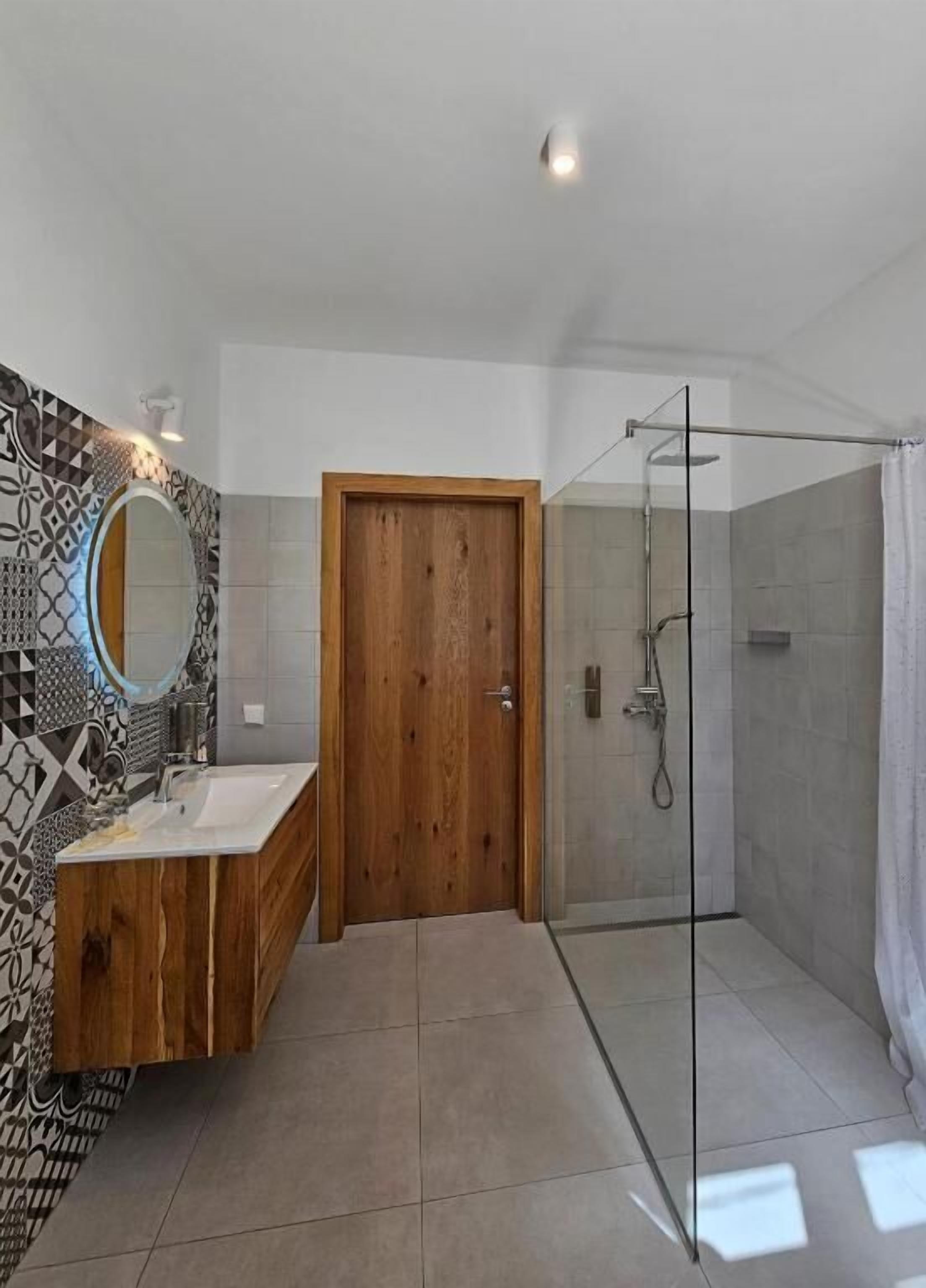 A modern bathroom with a glass shower enclosure, a wooden door, and a tiled floor.