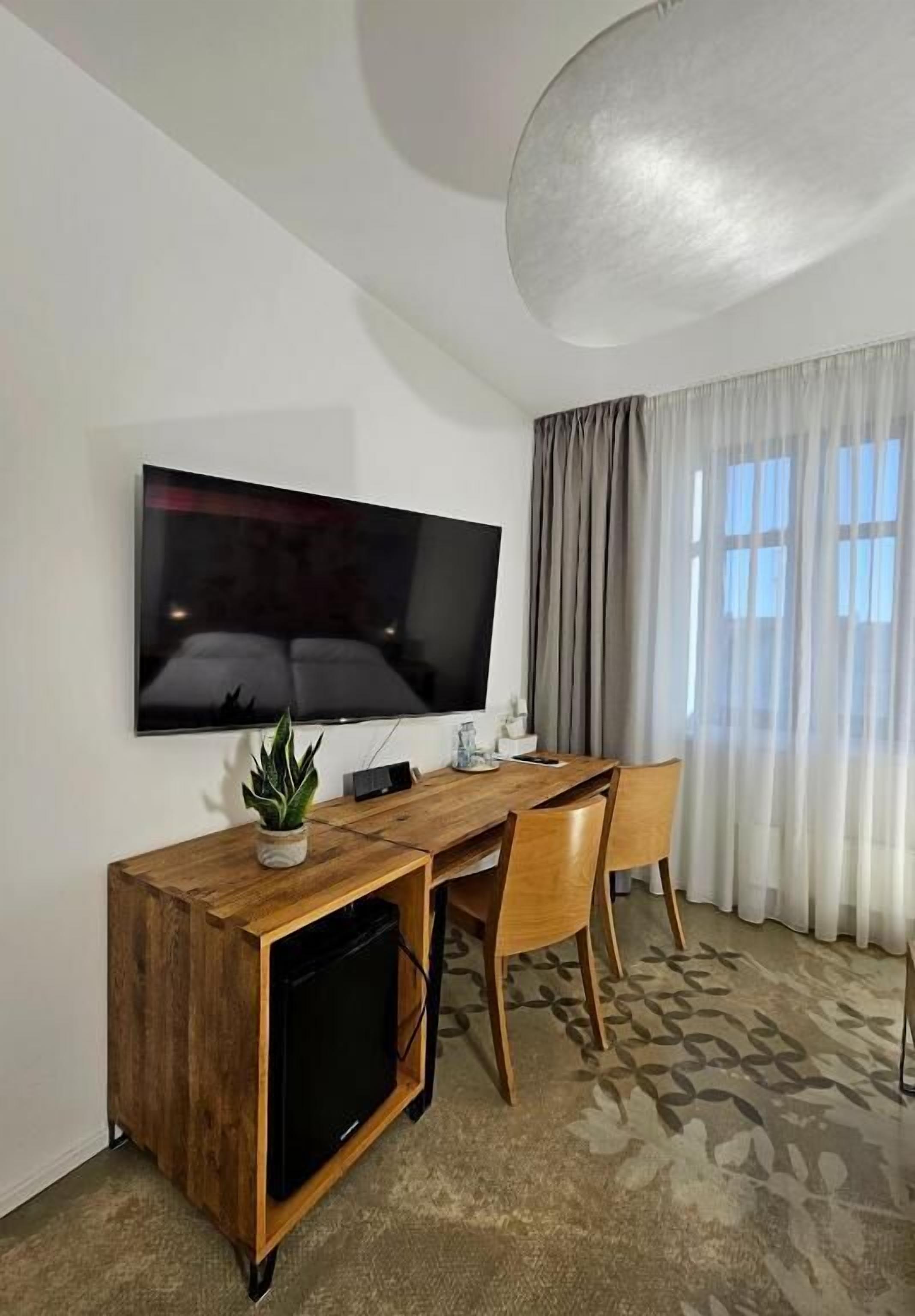 A hotel room with a flat-screen TV mounted on the wall, a wooden desk with a chair, a small plant, and a carpeted floor.