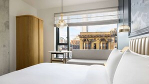 Ett hotellrum med en säng, ett litet bord, en stol, ett fönster med utsikt över byggnader och en hängande lampa.
