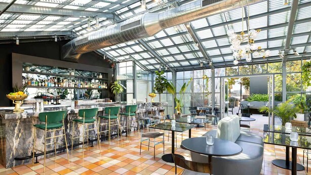 Un espace bar moderne avec un plafond en verre, des tabourets de bar verts et une variété de bouteilles exposées sur des étagères.