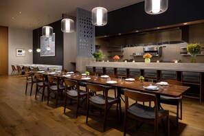 Un'area pranzo moderna con un lungo tavolo in legno apparecchiato per più persone, illuminazione a sospensione e una cucina sullo sfondo.