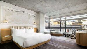 Ett modernt hotellrum med en stor säng, ett skrivbord, en stol och utsikt över staden genom stora fönster.