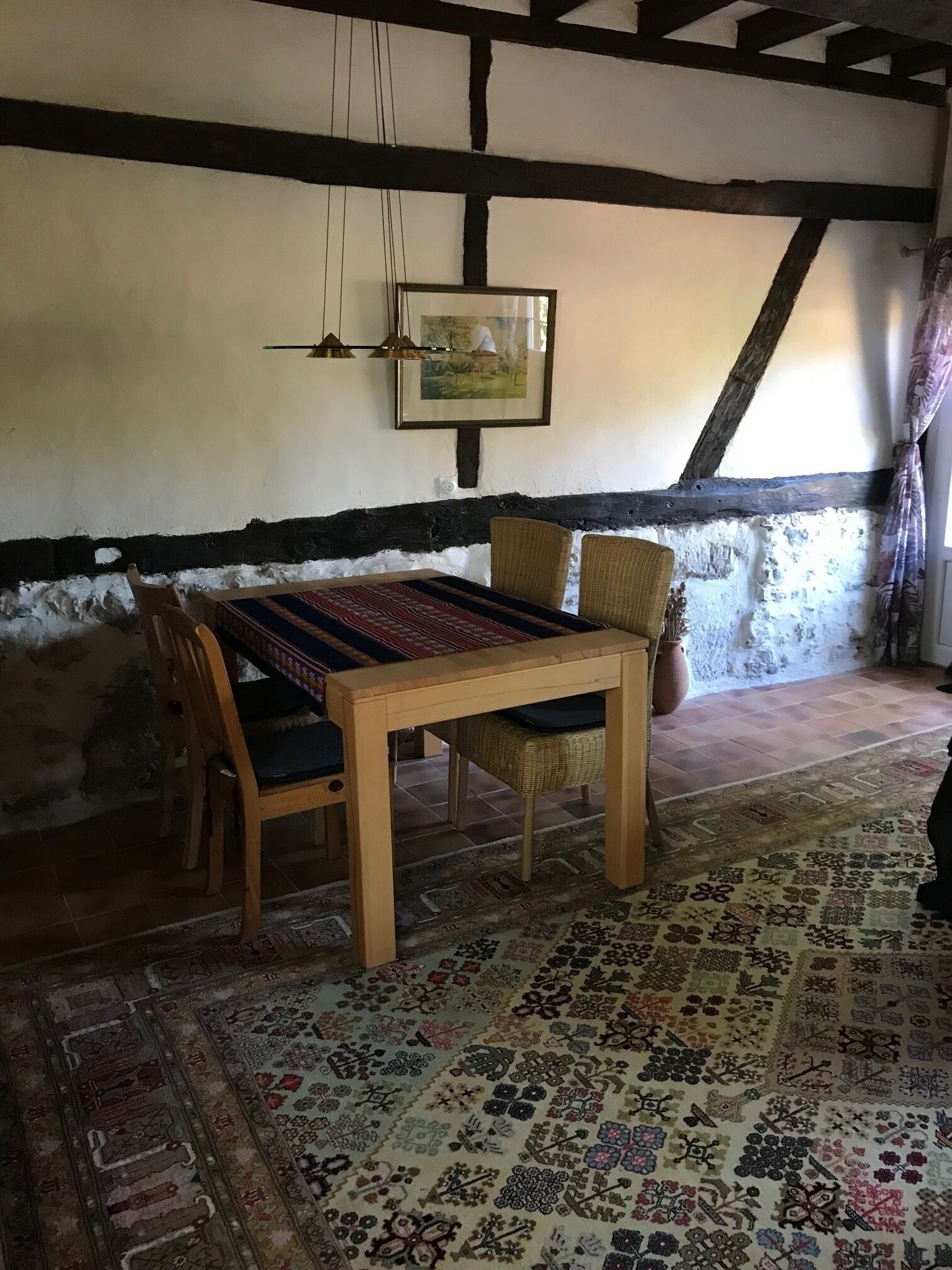 A rustic room with a wooden table, two wicker chairs, a patterned rug, a framed picture on the wall, and a sloped ceiling with exposed beams.