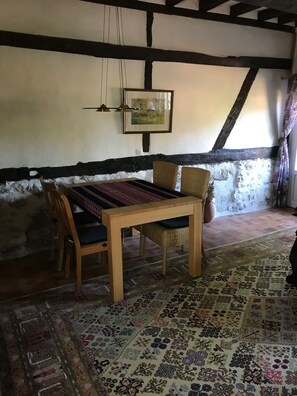 A rustic room with a wooden table, two wicker chairs, a patterned rug, a framed picture on the wall, and a sloped ceiling with exposed beams. - The Enchanted Cottage (Saint-Christophe-sur-Condé)