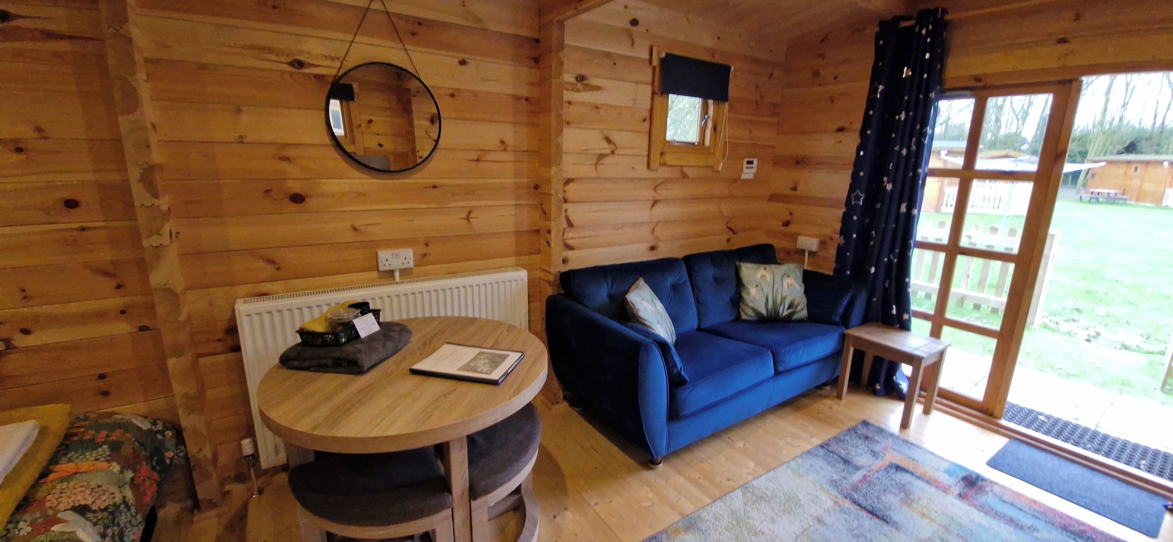 Una habitación acogedora con detalles de madera, una mesa redonda con un libro, un sofá azul con cojines y una ventana con cortinas.