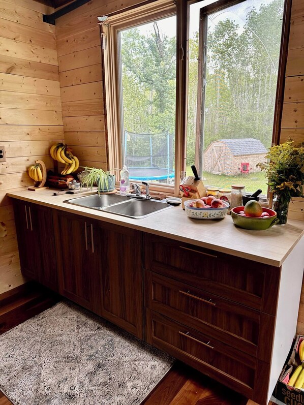 A wooden cabin kitchen with a large window, wooden cabinets, a sink, and various kitchen items.