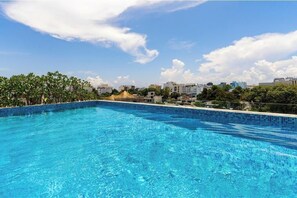 En klarblå swimmingpool med en kant av kakel, omgiven av grönska och byggnader i bakgrunden.