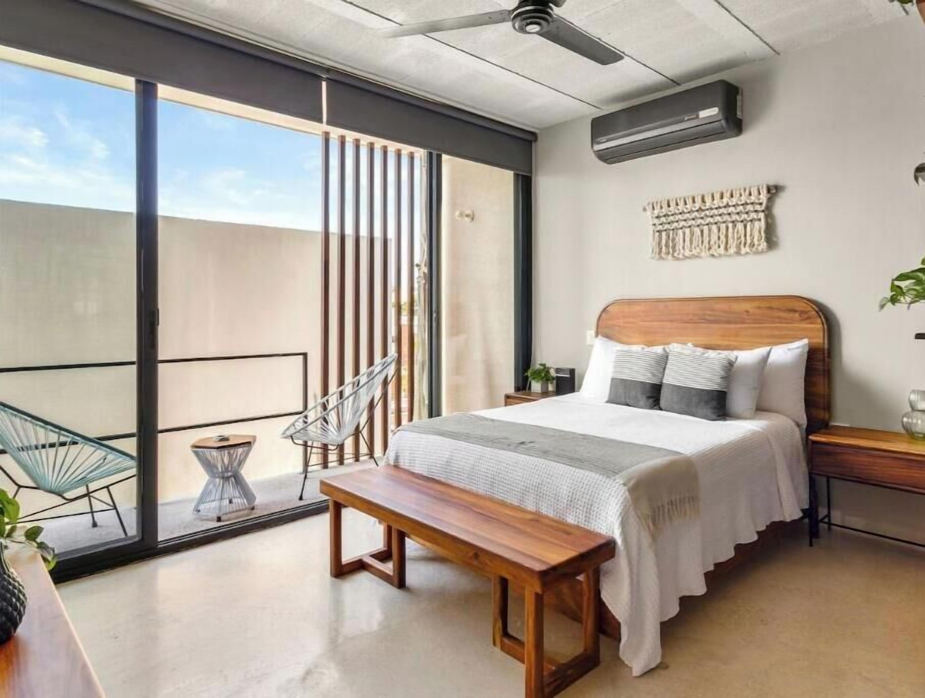 A modern bedroom with a large sliding glass door leading to a balcony, a wooden bed with a headboard, a bench, and a small side table with a plant.