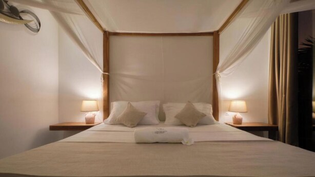 A bed with a canopy, two bedside lamps, and a pillow with a tag. - LE KEPARANGA (Mbour)