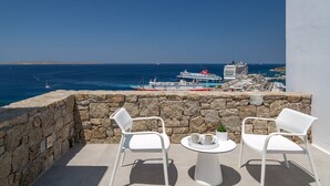 A balcony with stone walls, two white chairs, a small round table, and a potted plant. In the background, there is a view of the sea with boats and a cruise ship docked at a port. - Villa Corenyth, Divine Sea Views, Private Furnished Terrace, Shared Pool,. (Mykonos)