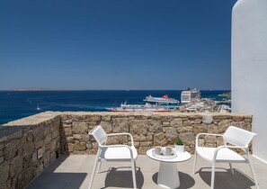 Ein Balkon mit Steinwänden, zwei weiße Stühle, ein kleiner runder Tisch und ein Blumentopf. Im Hintergrund ist ein Meerblick mit Booten und einem Kreuzfahrtschiff zu sehen, das im Hafen liegt.