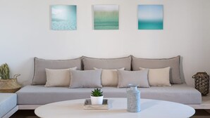 A living room with a grey sofa, three wall-mounted paintings, a white coffee table with a plant and a vase, and a wicker basket. - Villa Corenyth, Divine Sea Views, Private Furnished Terrace, Shared Pool,. (Mykonos)