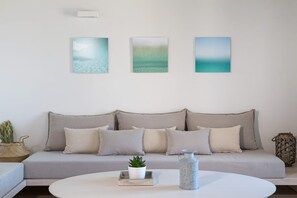 Ein Wohnzimmer mit grauem Sofa, drei Wandbildern, einem weißen Couchtisch mit Pflanze und Vase sowie einem Korbwäscher.