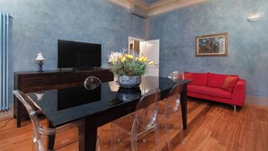 Een kamer met een eettafel, stoelen, een rode bank, een televisie en een vaas met bloemen.