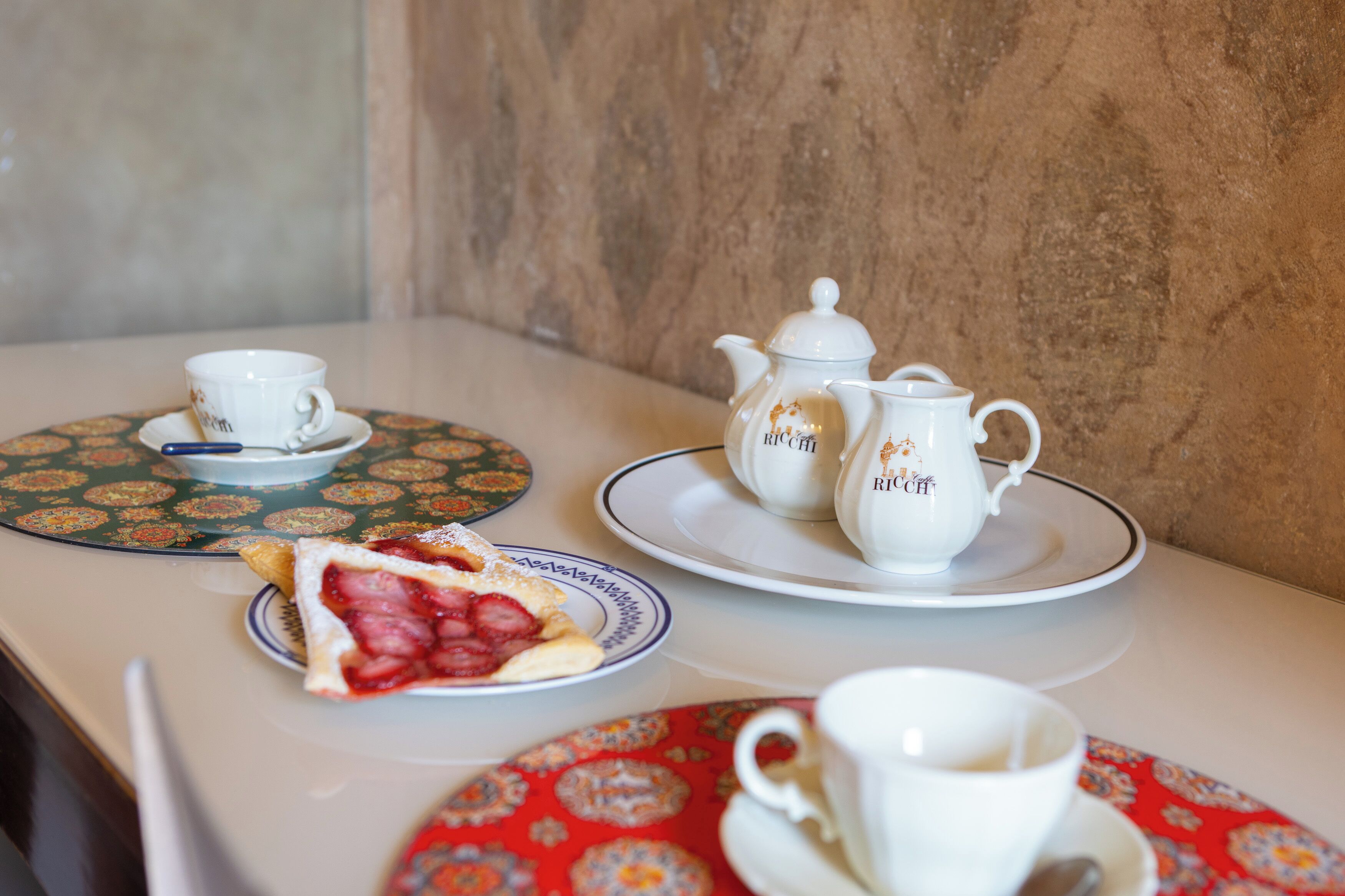 A table with a teapot, cups, saucers, and a slice of strawberry shortcake.