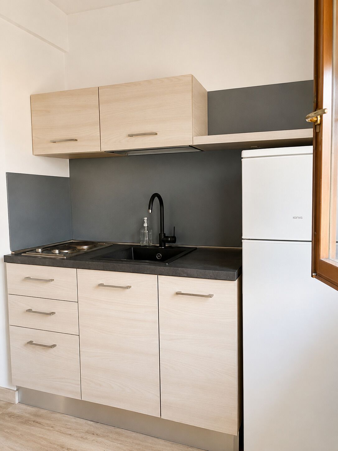 Une cuisine de taille réduite, avec des meubles de couleur bois clair, un évier noir et un réfrigérateur blanc.