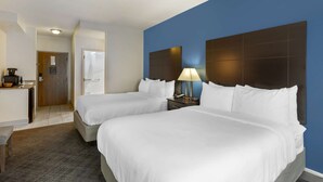 A hotel room with two beds, a desk with a coffee maker, a lamp, and a door leading to a bathroom.