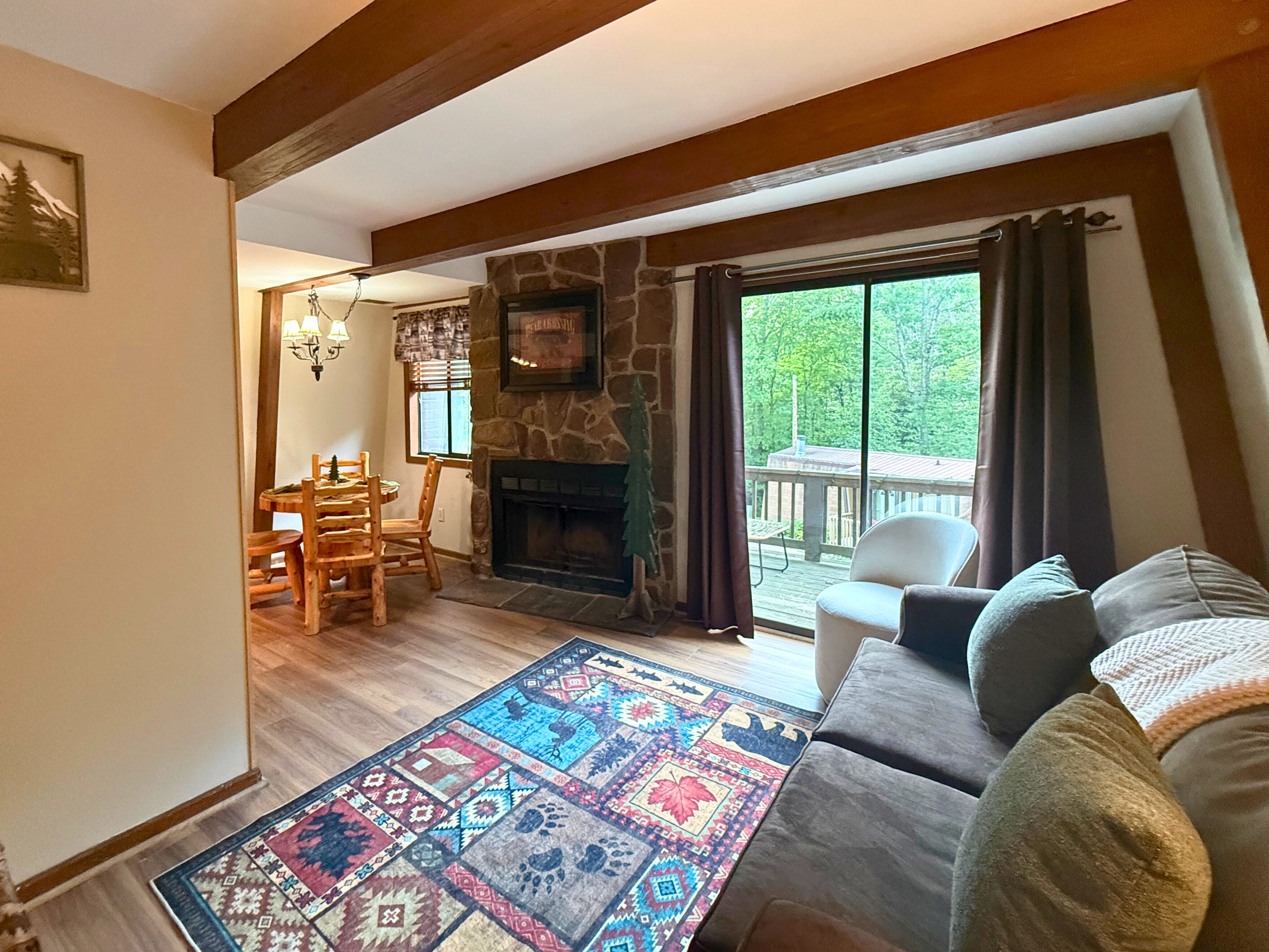 A living room with a stone fireplace, a wooden dining table with chairs, a colorful rug, and a view of a deck through a large window.