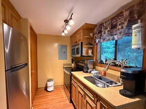 A kitchen with a stainless steel refrigerator, microwave, and coffee maker, wooden cabinets, a window with a view, and a trash can.