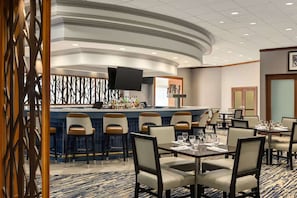 Um bar de hotel com balcão curvo, banquetas e uma tela de televisão. Há mesas de jantar com cadeiras dispostas no espaço.
