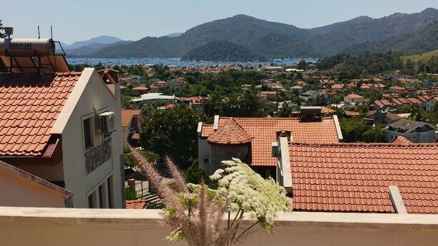 A balcony with a vase of flowers, a white tablecloth, and a view of a coastal town with red-roofed buildings and mountains in the distance. - Stunning Private Panorama Suite within a 350m² Luxury Villa – Full Bay View" (Muğla)