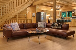 A log cabin interior with a brown leather sectional sofa, a round coffee table, and a kitchen area with wooden cabinets and a stainless steel refrigerator. - Misty Mountain River House*Fire Pit*Private Cabin (Tillamook)