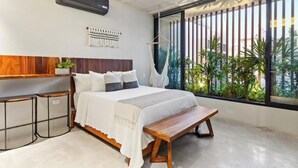 A modern bedroom with a large bed, bedside tables, a hanging chair, and a view of greenery through large windows. - Ocean Boutique Condos (Playa del Carmen)