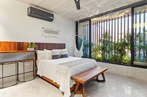 Ein modernes Schlafzimmer mit einem großen Bett, Nachttischen, einem Hängekorbstuhl und Blick auf Grün durch große Fenster.