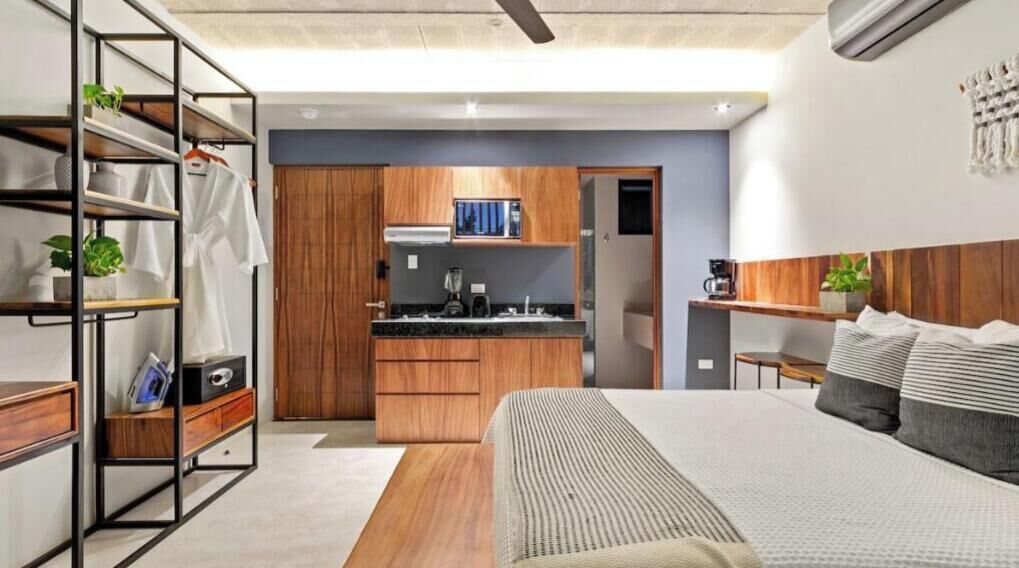 Un dormitorio moderno con una cama grande, un armario de madera, una cocina integrada y una estantería con plantas.