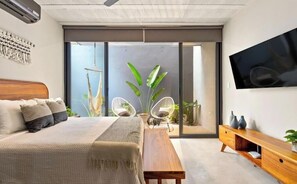 Ein modernes Schlafzimmer mit einem großen Bett, einer Holzbank, zwei weißen Korbstühlen, einem Fernseher an der Wand und einer Glastür zum Balkon mit Pflanzen.