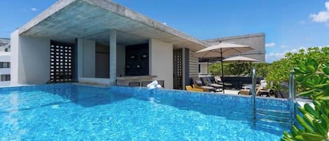 A swimming pool with lounge chairs and umbrellas, a covered seating area, and a building with a modern architectural style.