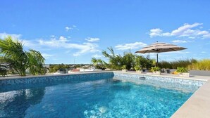 Una piscina blava i cristal·lina amb vora de rajola, envoltada de verdura i un cel assolit.