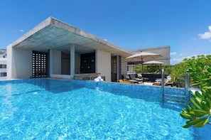 Ein Schwimmbecken mit Liegestühlen und Sonnenschirmen, eine überdachte Sitzgelegenheit sowie ein Gebäude im modernen architektonischen Stil.