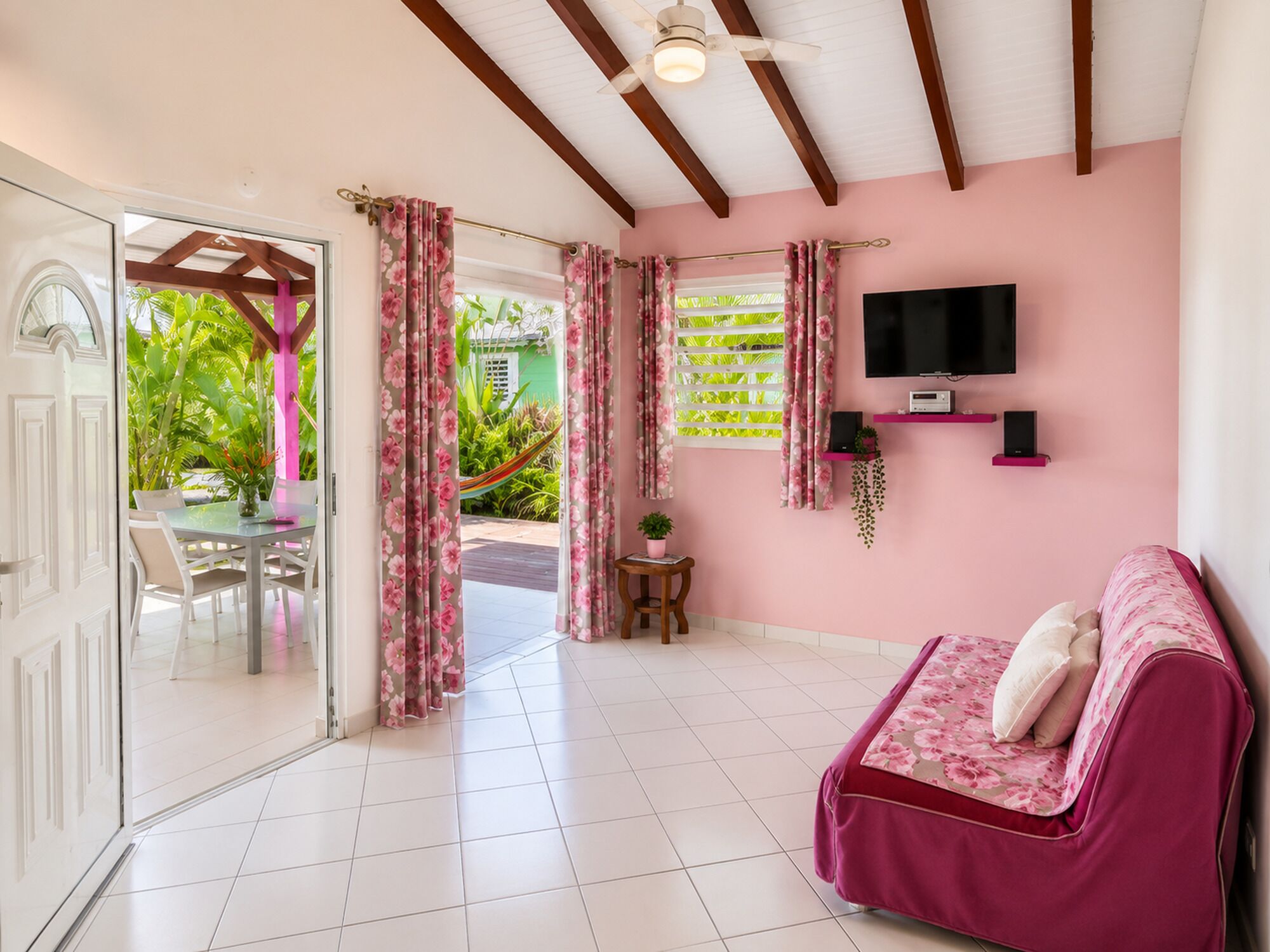 Ein Zimmer mit rosafarbenen Wänden, einem Bett mit floralem Bettzeug, einem Wandfernseher und einem Fenster mit Vorhängen.