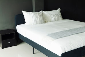 A neatly made bed with white bedding and patterned pillows, a dark blue headboard, and a black nightstand with a drawer. - The One Rawai Rooms (Rawai)