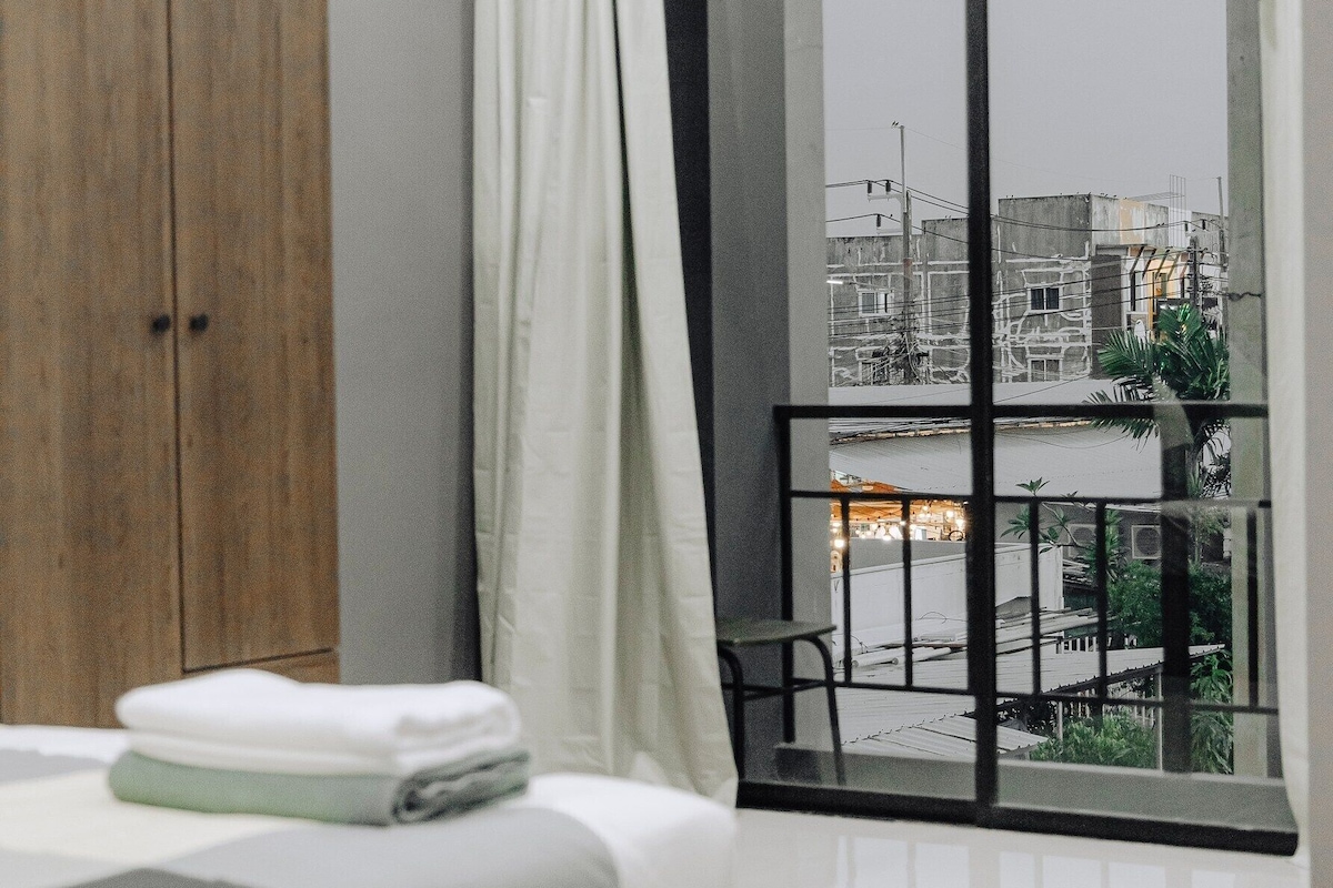 A modern bedroom with a bed, a wooden wardrobe, a window with a view of buildings, and an air conditioning unit.
