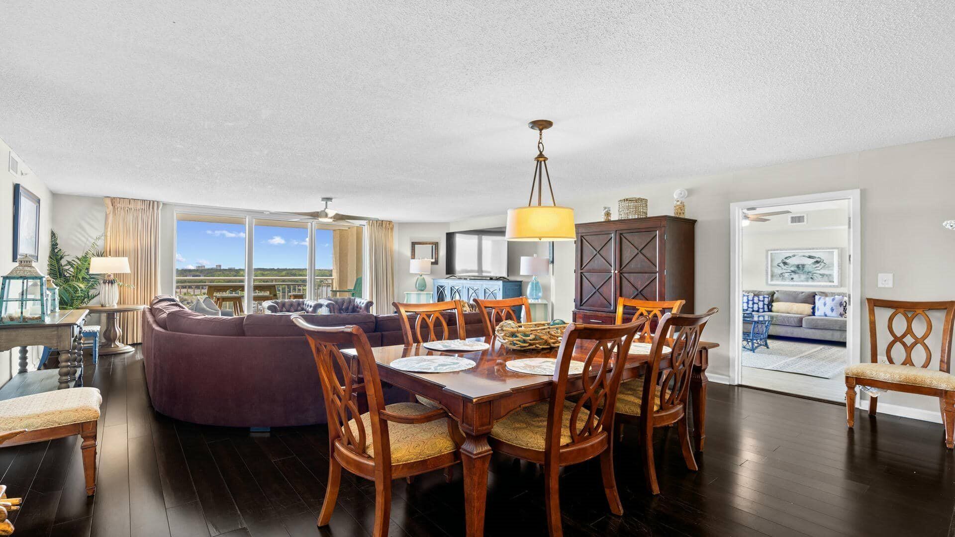 Un salón espacioso con suelo de madera oscura, un sofá morado, una mesa de comedor con sillas y un ventanal con vistas.