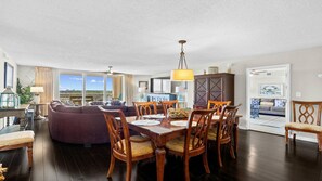 A spacious living area with a dark wood floor, a purple sofa, a dining table with chairs, and a large window offering a view.